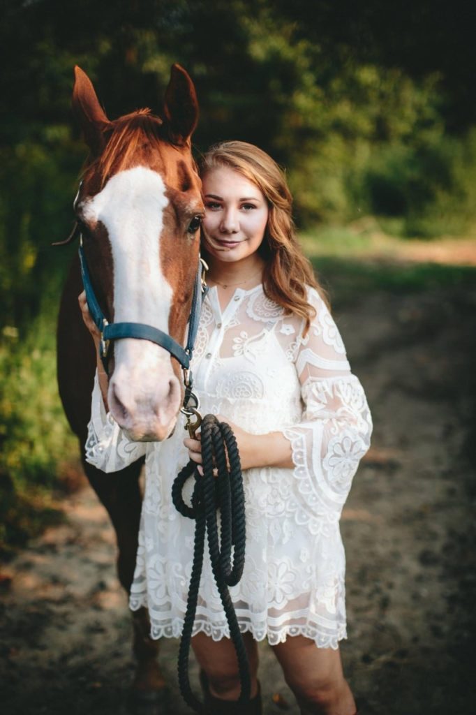 Gabby, a white woman with medium length curly blonde hair, is standing next to a chesnut horse.