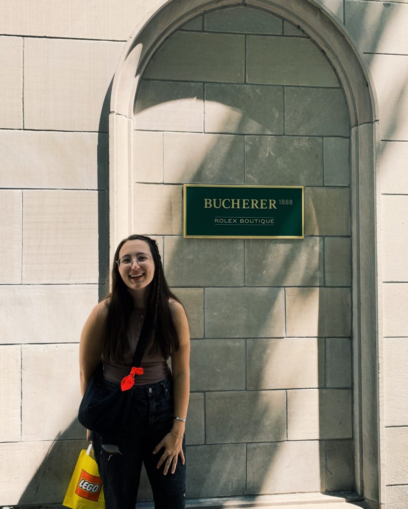 A woman with long dark hair holing a Lego bag and standing in front of a Rolex plaque on a stone wall