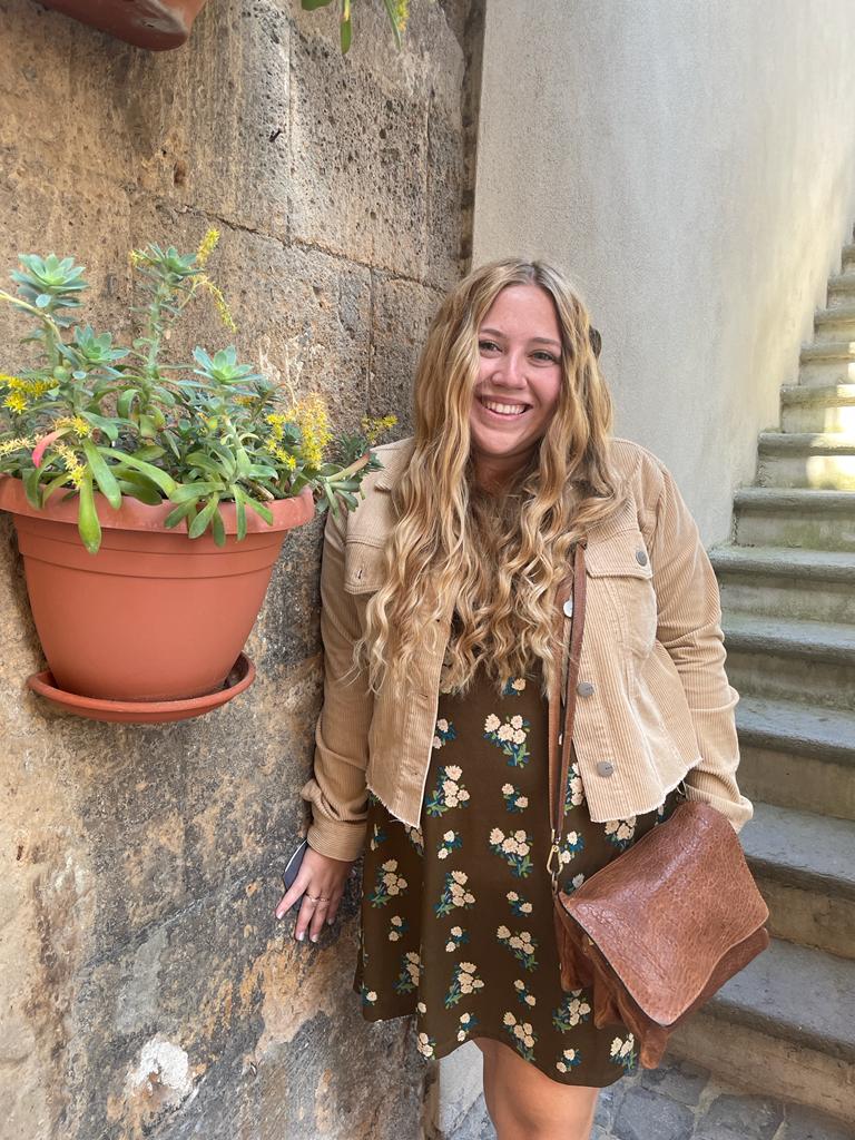 Katie, a white woman with long, wavy blonde hair smiling next to a plant wall in Italy
