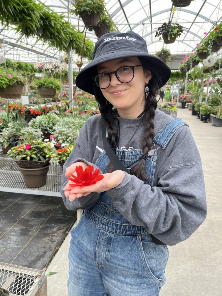 Julia, a person with a dark brown hair holding a red flower up towards their face and smiling.