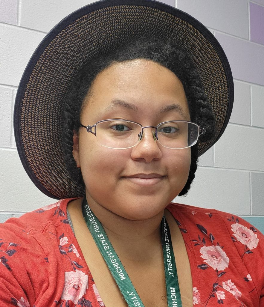 Raven, a smiling woman with dark hair, glasses, a straw hat, a green MSU lanyard in a red shirt.