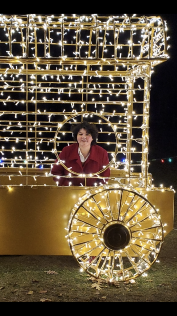 Oliviah, a woman wearing a red coat surrounded by Christmas lights, smiling through a round window.