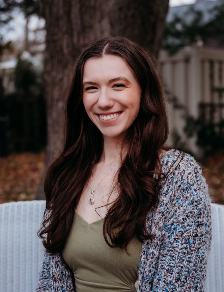 Emily, a white woman with long brown hair smiling in front of a tree sitting on a white wicker couch