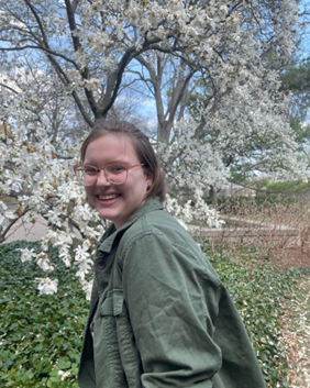 Image of a smiling white woman with glasses, short brown hair, and a green jacket. In the background, there is a flowery tree and foliage.