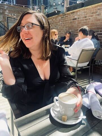 A femme white person—with brunette hair, glasses, and a black dress—smiles as they drink her coffee.